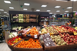 Ein Geschäft mit Körben voller Obst und Gemüse, Preisschilder, einem geschmückten Weihnachtsbaum links und Regalen mit anderen Artikeln im Hintergrund.