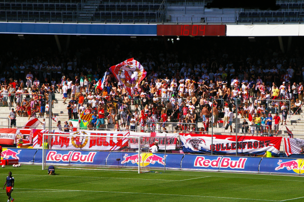Ein Fuballspiel in einem Stadion mit Spielern auf dem Feld, einem Tor mit Netz, Bannern, einem Metallzaun, einer Anzeigetafel, einem Display und einem Dach mit Deckenbeleuchtung.