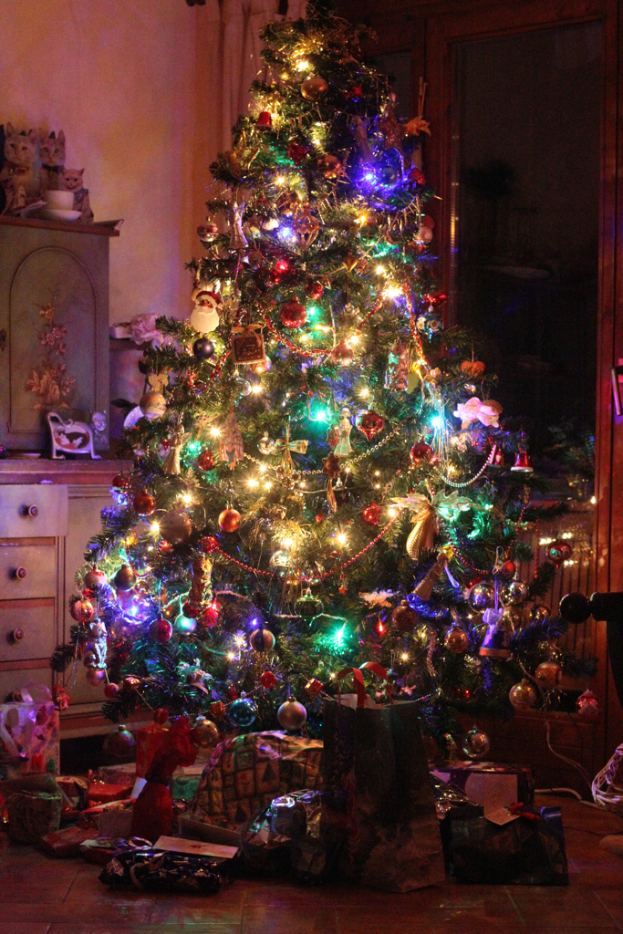 Ein Weihnachtsbaum mit Lichtern und Deko, Geschenke auf dem Boden, ein Schrank mit Gegenst├Ąnden links und ein Fenster an der rechten Wand.
