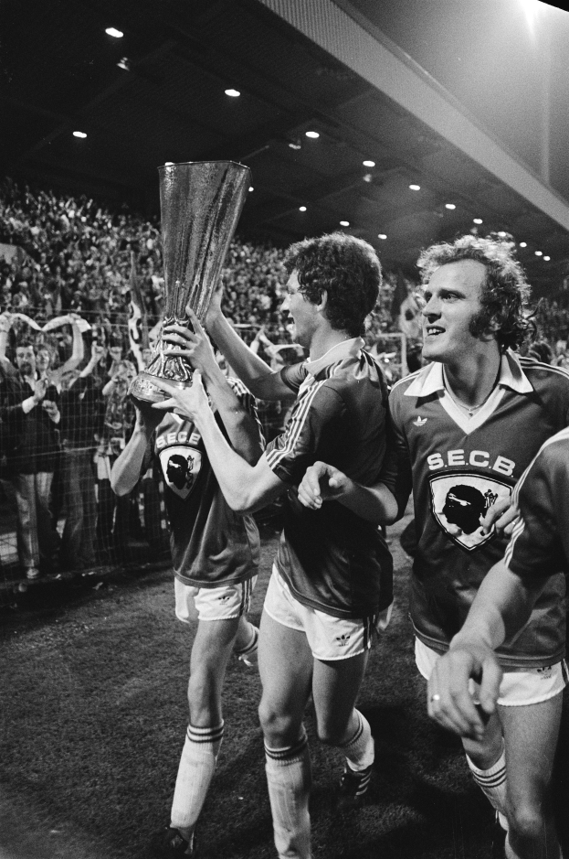 Eine Gruppe von Männern in schwarzer und weißer Kleidung hält einen Pokal auf einem Fußballfeld hoch, während sie ihren FA-Cup-Finalsieg feiern, umgeben von einer Menge hinter einem Zaun unter Stadionbeleuchtung.