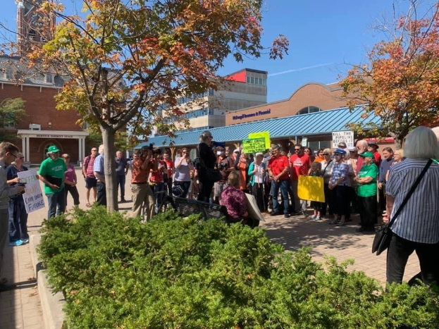 Eine Gruppe von Studenten demonstriert vor dem Campus der University of Michigan, mit Schildern und einigen mit Kameras, mit Bäumen und Himmel im Hintergrund.