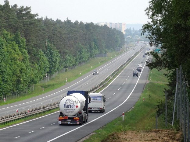 Ein Lkw fährt auf einer Autobahn mit Bäumen auf beiden Seiten, einem Zaun und Gras auf der rechten Seite, Gebäuden im Hintergrund und einem klaren blauen Himmel.