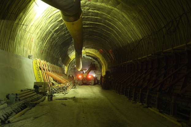 Baustelle mit einem großen Tunnel, Fahrzeugen, verstreuten Holzobjekten, Rohren, einer Wand auf der linken Seite und Deckenbeleuchtung.
