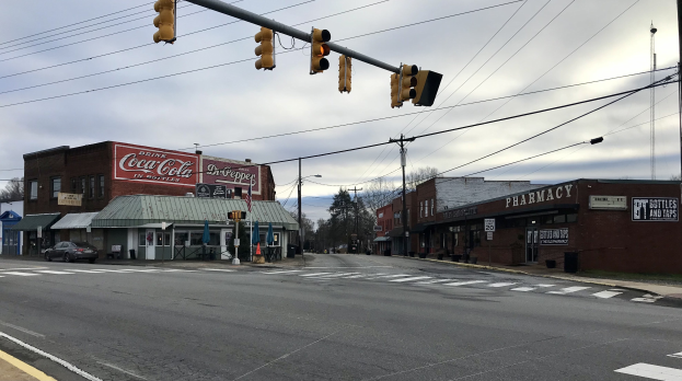 Stadtstraßenkreuzung mit Ampel, Fahrzeugen, Gebäuden mit Schildern, Strommasten mit Kabeln, Bäumen und bewölktem Himmel.