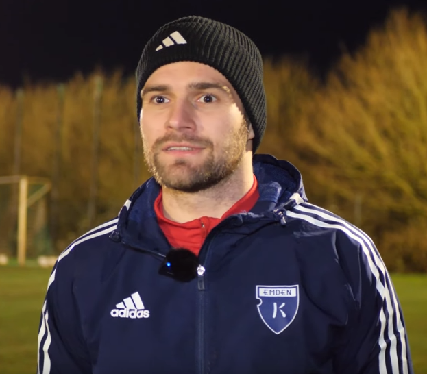 Ein Mann in einer blauen Jacke und Mütze steht auf einem Fussballfeld, umgeben von Bäumen und einem Tor im Hintergrund, während eines Trainings.
