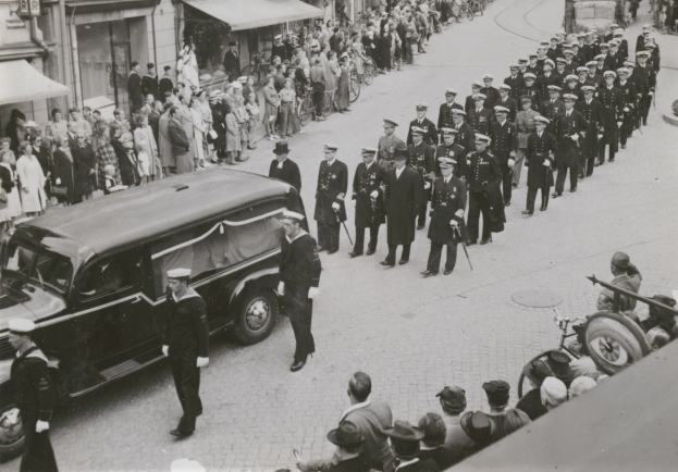 Schwarz-weißes Bild einer Trauerprozession mit einem Leichenwagen, der den Leichnam eines Mannes trägt, umgeben von Menschen mit Waffen und Fahrzeugen auf der Stra├če, mit Geb├Ąuden und Schildern im Hintergrund.