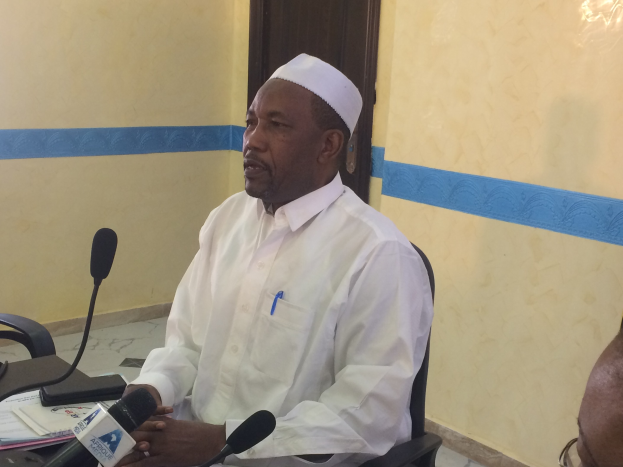 Ein Mann in einem weißen Gewand und Hut sitzt an einem Tisch mit Mikrofonen, während er spricht und Papiere und Gegenstände auf dem Tisch liegen, mit einer anderen Person zu seiner Rechten und einer Tür im Hintergrund.