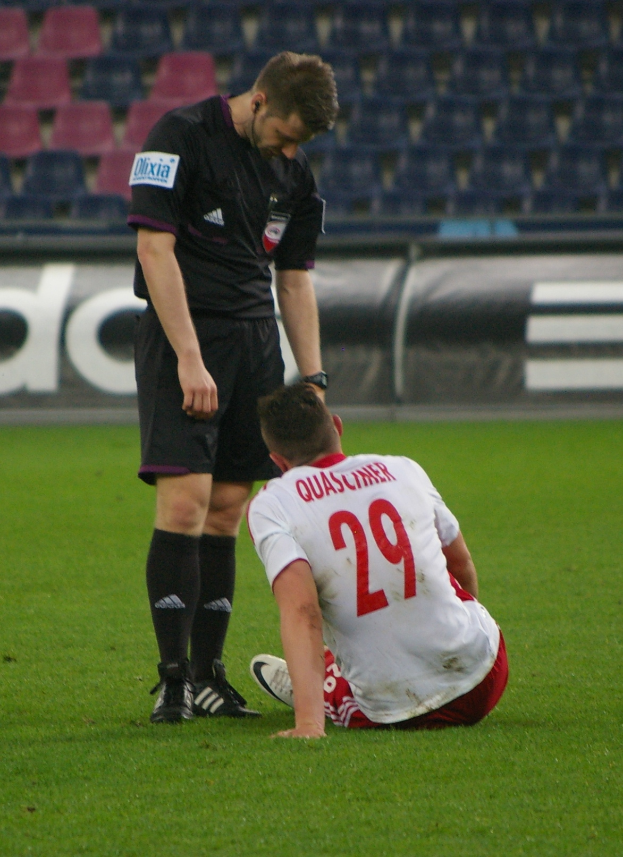 Ein Fußballspieler sitzt neben einem Schiedsrichter auf dem Boden in einem Stadion, beide tragen Sportbekleidung, mit Tafeln und Stühlen im Hintergrund.