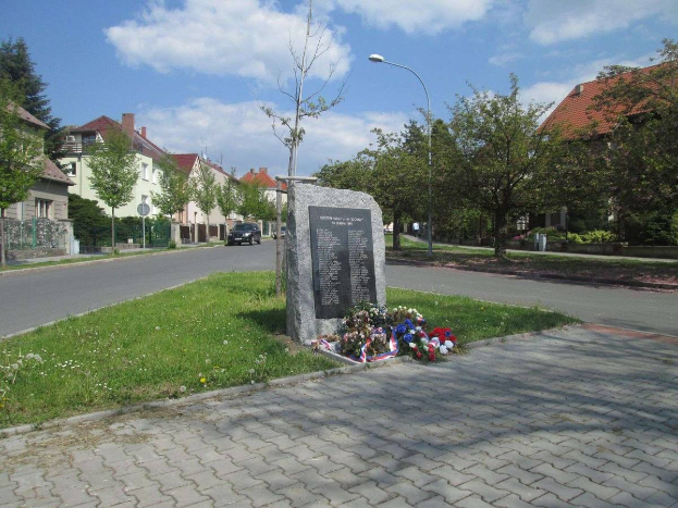Ein Gedenkstein in einer Stra{\u}enmitte umgeben von Gras, Pflanzen, B"{a}umen, Laternenmasten und Fahrzeugen, mit Geb"{a}uden und einem bew"{o}lktem Himmel im Hintergrund, gewidmet den Opfern des Holocaust.