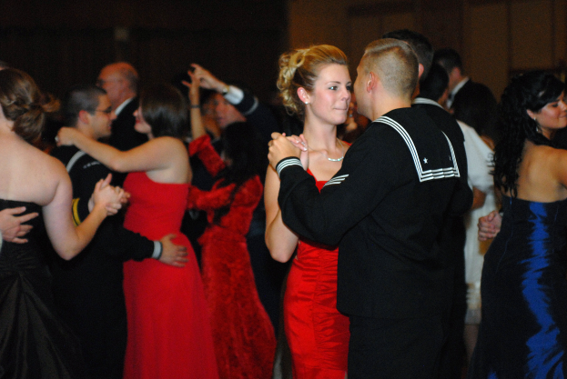 Eine Gruppe von Menschen in eleganter Kleidung tanzt energisch auf einer Tanzfläche mit einer Wand im Hintergrund.