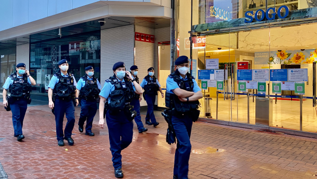 Eine Gruppe von Polizisten in Masken geht eine Straße entlang vor einem gläsernen Gebäude, mit einem Geschäft auf der rechten Seite, das Plakate auf seinen Glaswänden zeigt.