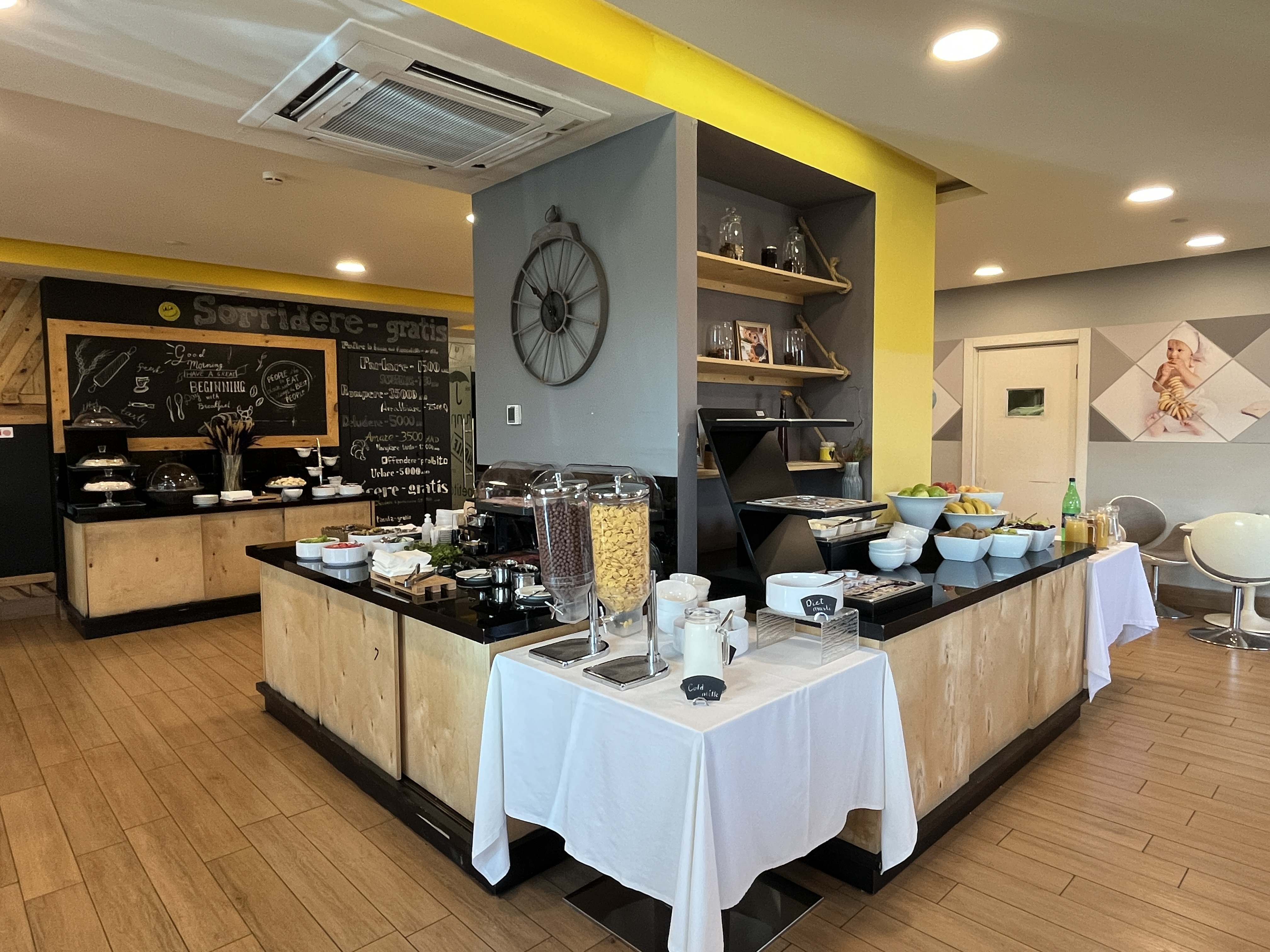 Ein Restaurantinnenraum mit Tischen und Stühlen, Essensbehältern, einer Tafel mit Text, einer Wanduhr, Deckenleuchten, einer Klimaanlage und einer Tür im Hintergrund, das sich im Hôtel de Ville in Paris befindet.