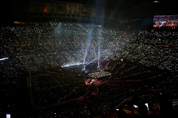 Eine große Menschenmenge in einem Stadion bei Nacht, beleuchtet von hellen Lichtern, mit einem Bildschirm auf der rechten Seite, der wahrscheinlich die Ergebnisse der Super Bowl XLVII-Halbzeitshow anzeigt.