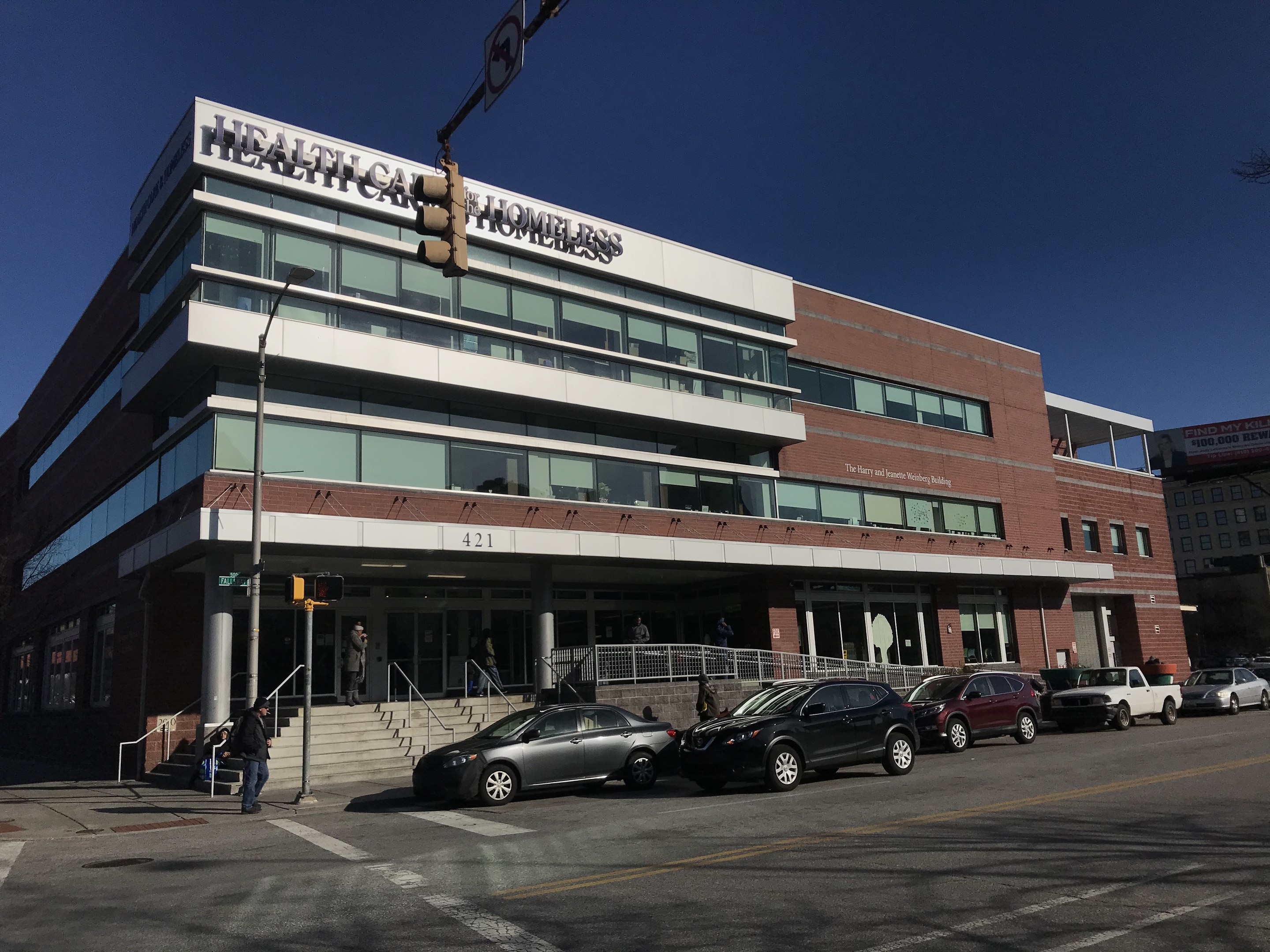 Großes Gebäude mit der Aufschrift "New York City Health Care Homeless Center" mit geparkten Autos, Passanten, Straßenmöblierung und Bäumen unter einem klaren blauen Himmel.