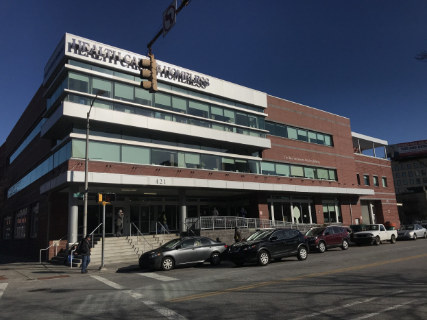 Großes Gebäude mit der Aufschrift "New York City Health Care Homeless Center" mit geparkten Autos, Passanten, Straßenmöblierung und Bäumen unter einem klaren blauen Himmel.