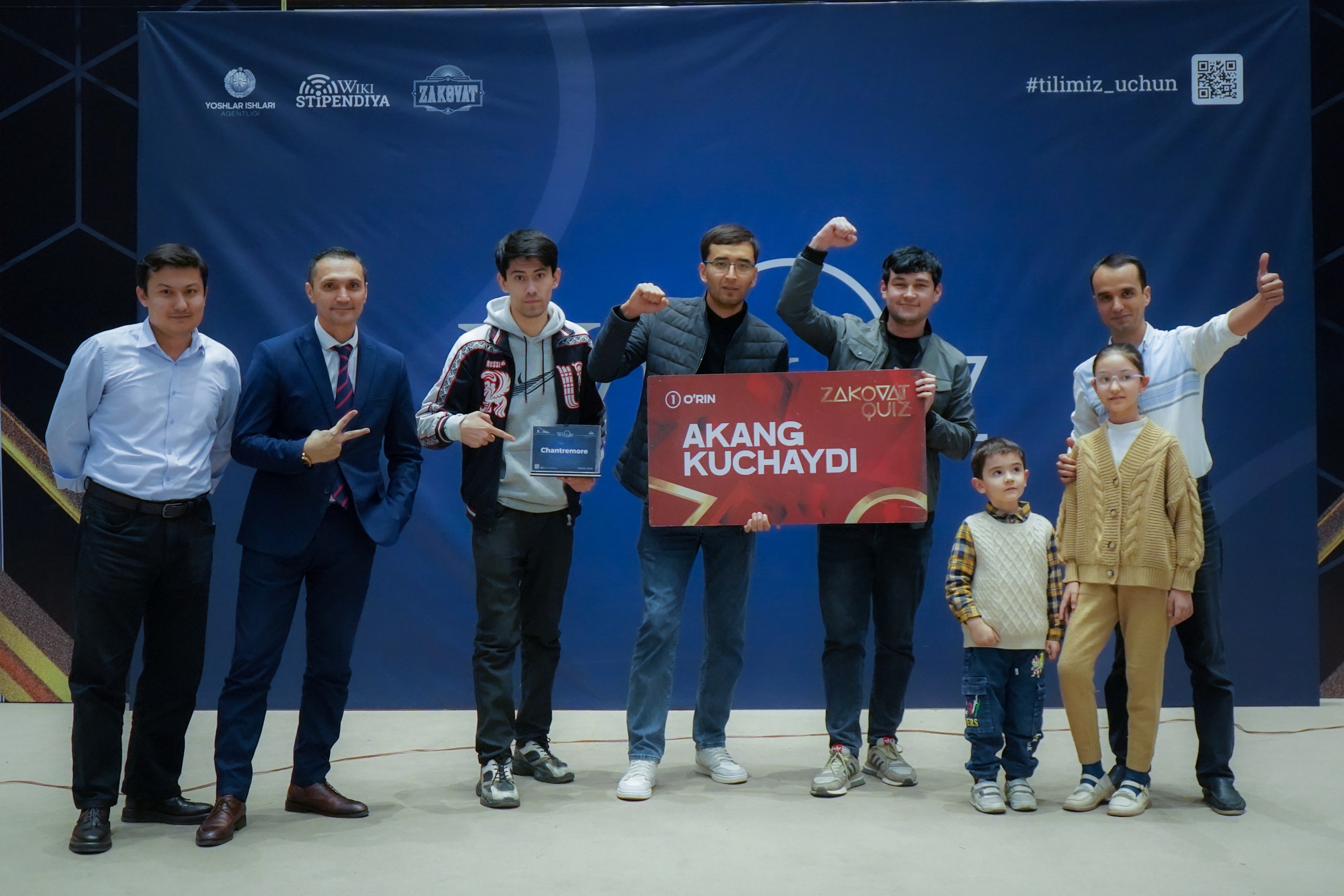 Gruppe von Menschen auf einer Bühne mit einer Tafel, auf der "Akang Kuchaydi - Zakovat Quiz" steht, mit einem Banner, Lichtern und anderen Gegenständen im Hintergrund.