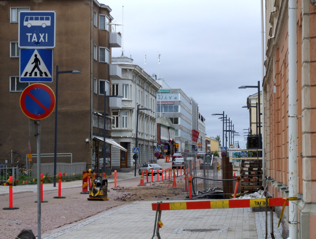 Eine Stadtstraße mit Gebäuden, Straßenlaternen, Verkehrsschildern, Verkehrsleitkegeln, Kraftfahrzeugen, Absperrpollern, Bäumen und einer Baustelle mit Verkehrszeichen unter einem bewölkten Himmel.