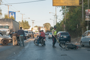 Eine Gruppe von Menschen steht um ein verunglücktes Motorrad auf der Seite einer Straße mit mehreren Fahrzeugen, darunter ein Lastwagen, und Hintergrundelementen wie Bäumen, Mästen, Lichtern, Schildern und dem Himmel.