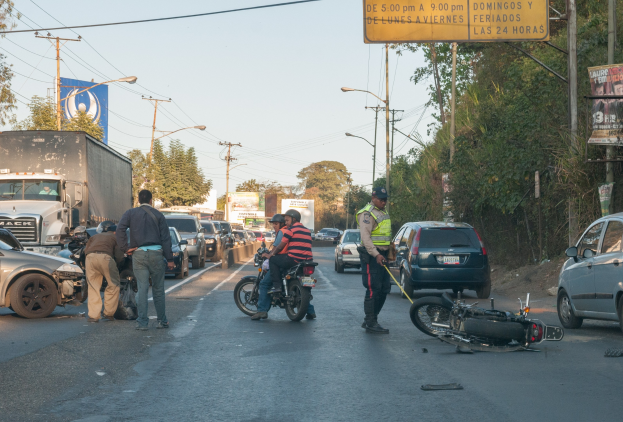 Eine Gruppe von Menschen steht um ein verunglücktes Motorrad auf der Seite einer Straße mit mehreren Fahrzeugen, darunter ein Lastwagen, und Hintergrundelementen wie Bäumen, Mästen, Lichtern, Schildern und dem Himmel.