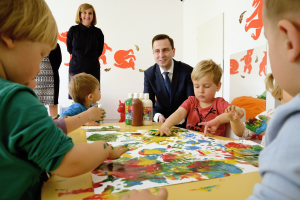 Kinder sitzen um einen Tisch mit Farbe an den Händen, umgeben von Papieren und Flaschen, während eine Frau im Hintergrund lächelt, umgeben von Gemälden an der Wand.