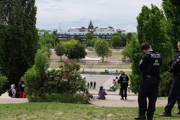 Zwei Polizeibeamte vor einer Gruppe von Menschen in einem Park mit grünem Gras, Bäumen, bunten Blumen, Gebäuden, Polen und einem klaren blauen Himmel.