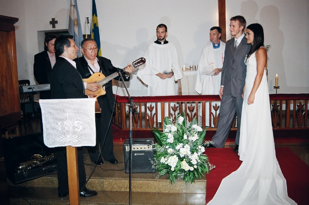 Eine HochzeitsSzene in einem Raum mit zwei Pastoren, einer Braut, einem Bräutigam und drei Musikern; ein Podium mit Stoff vor den Musikern; ein Tisch mit Kerzen hinter den Pastoren; eine Kerze in einem Halter hinter dem Paar; ein Strauß, Lautsprecher, Taschen und ein roter Teppich auf dem Boden; und Flaggen, eine Wand mit einem Kreuz, einen Schrank und einen Stuhl im Hintergrund.