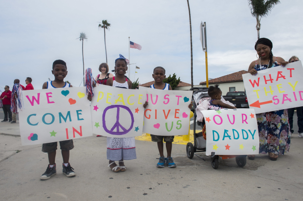 Eine Gruppe von Kindern hält Schilder und steht vor einem Gebäude, während eine Frau einen Kinderwagen mit einem Baby schiebt; auf einem Schild steht 'Wir kommen in Frieden, gebt uns unseren Daddy.'