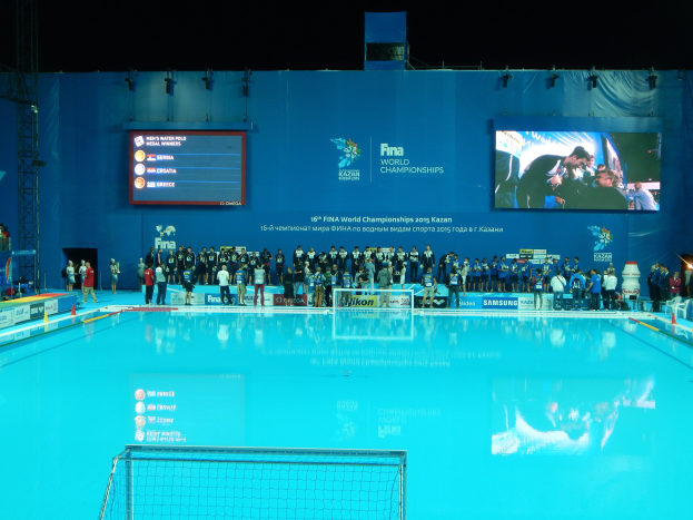 Gruppe von Menschen, die um ein Hallenbad mit Netz am Boden stehen, mit einer Wand im Hintergrund, die "FINA World Championships 2015 Kazan"-Text und Logos anzeigt, sowie Gerüsten und Lichtern.