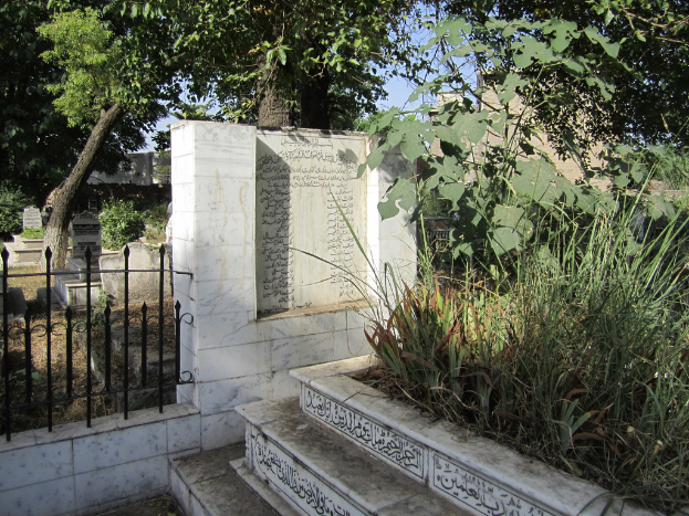 Ein Blick auf einen Friedhof mit Treppe unten, einer Gedenktafel in der Mitte, Gräbern hinter einem Geländer, Gras und Bäumen rechts und Gebäuden im Hintergrund.