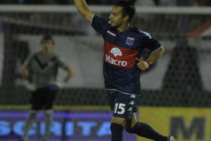 Ein Fußballspieler feiert nach einem Tor auf dem Boden, mit einer jubelnden Menge und einer Person im Hintergrund und einer Tafel mit Text dahinter, alles vor einem leicht unscharfen Hintergrund.