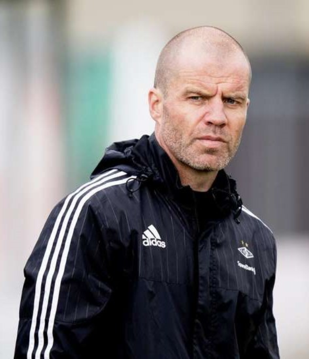 Bald coach in einer marineblauen Jacke mit weißen Streifen und weißem Kragen, der auf einem Fußballfeld steht und die Hände verschränkt hält