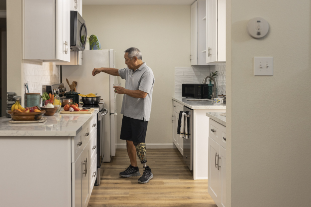 Ein Mann steht in einer Küche mit weißen Schränken, hält ein Glas in der Hand und ist von Obst, Gemüse, Schalen und Küchenutensilien auf einer Plattform zu seiner Linken umgeben, mit Geräten und Schränken im Hintergrund.